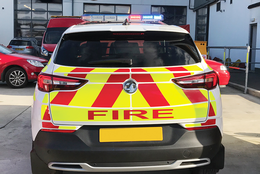 Back view of white fire response vehicle with partially lit red and blue lightbar on cemented vehicle forecourt in front of various vehicles and shutter doors